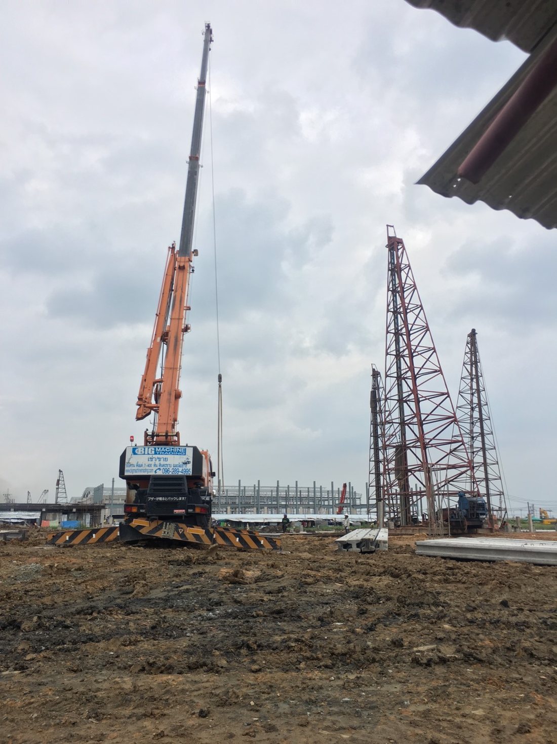 เครนล้อยาง BIG MACHINE TRADING สีส้มทำงานที่ไซต์ก่อสร้างศูนย์สมุนไพรไทย มีทาวเวอร์เครนสีแดงอยู่ใกล้เคียง