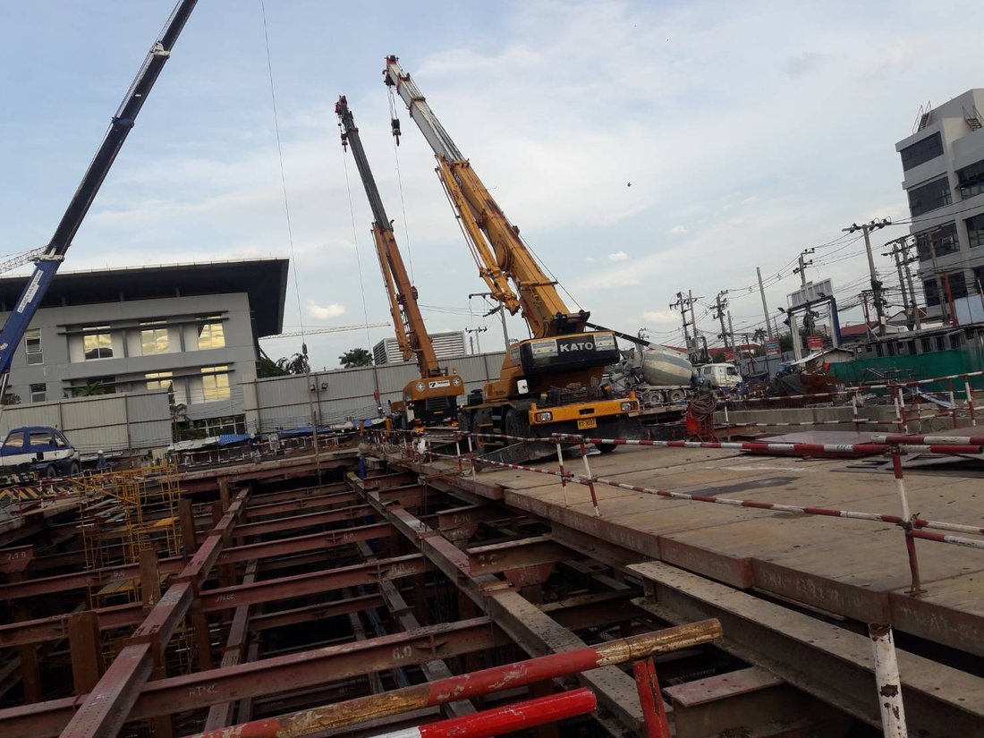 เครนล้อยาง KATO สีเหลืองของ BIG MACHINE TRADING จอดข้างเครนสีน้ำเงินที่ไซต์งานแจ้งวัฒนะ เห็นอาคารพักอาศัยด้านหลัง