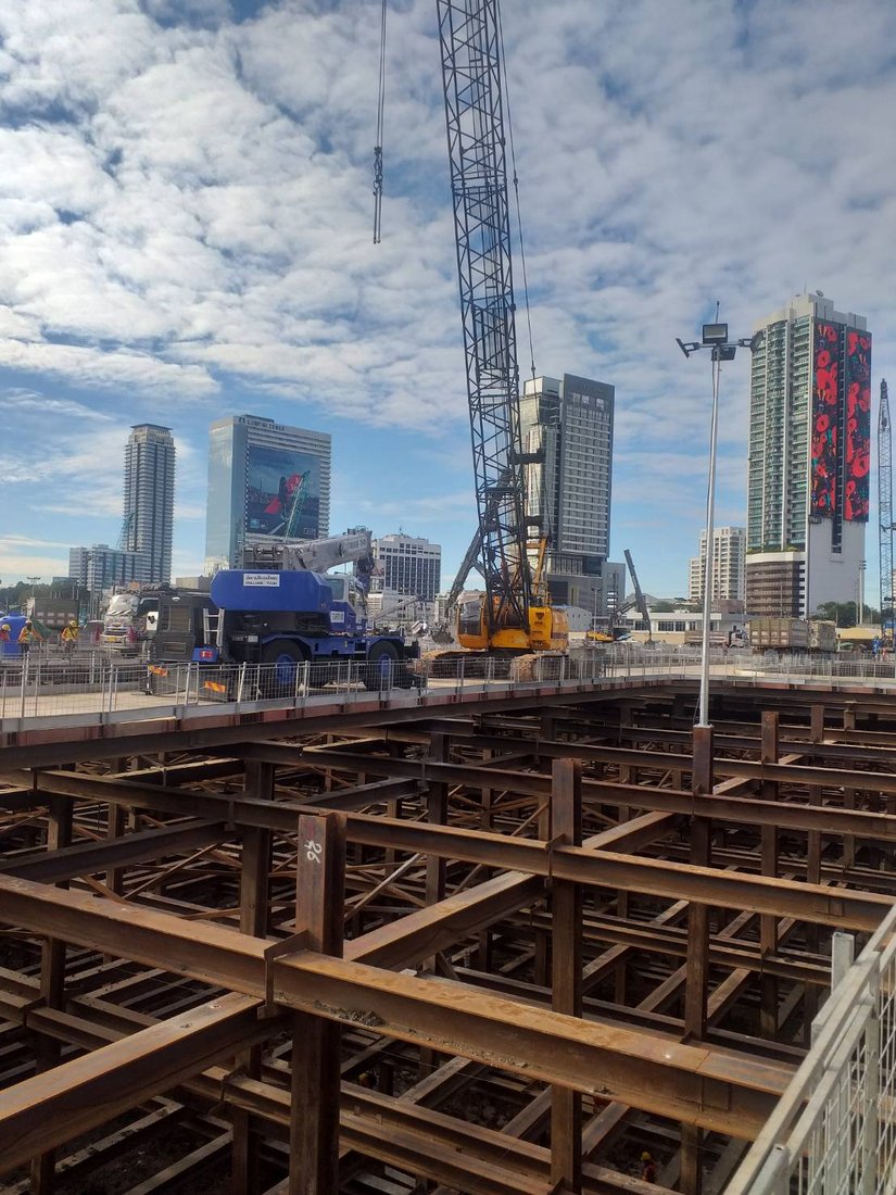 เครนล้อยางสีน้ำเงินและเครนตีนตะขาบของ BIG MACHINE TRADING ทำงานที่ไซต์ก่อสร้าง One Bangkok เห็นตึกสูงย่านสีลมด้านหลัง