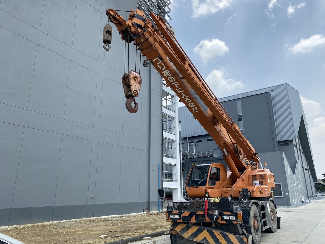 เครนล้อยาง BIG MACHINE TRADING ทำงานที่ไซต์ก่อสร้างโรงงานผลิตเวชภัณฑ์ ไทรน้อย นนทบุรี เห็นผนังอาคารสีเทาเข้มและโครงสร้างเหล็กด้านหลัง